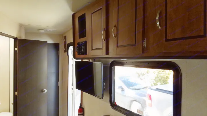 An RV rental interior features dark overhead cabinets, two TVs, a door, and a window showing parked vehicles.. Forest River Salem Cruise Lite 2017