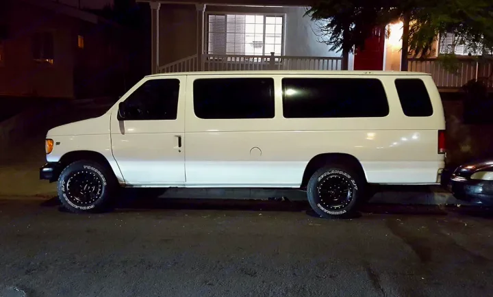 White Ford van with tinted windows and custom wheels, parked at night. Perfect for an RV rental adventure.. Ford E350 2000