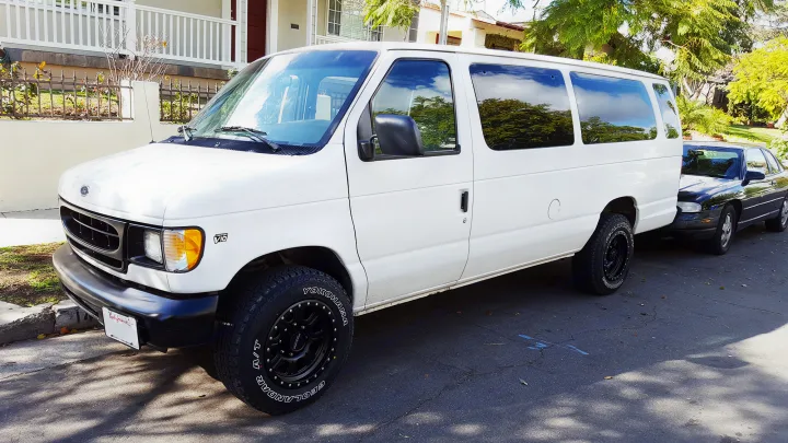 A white Ford V10 van with rugged tires, parked on a street in front of a black car. Could be a great rv rental.. Ford E350 2000