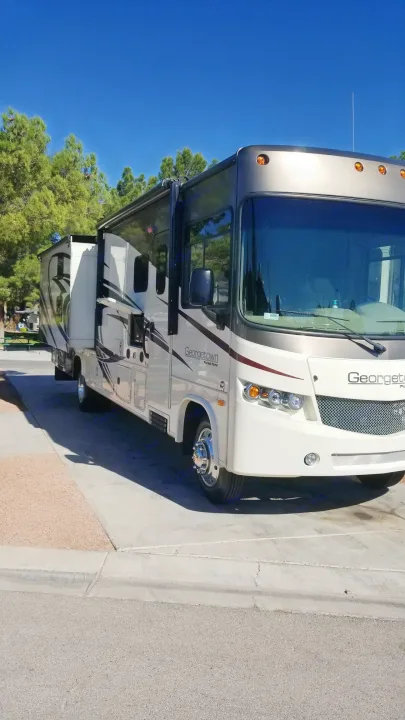 A modern Georgetown rv rental, a white and brown motorhome, is parked with slide-outs extended at a sunny campsite.. Forest River Georgetown 2017