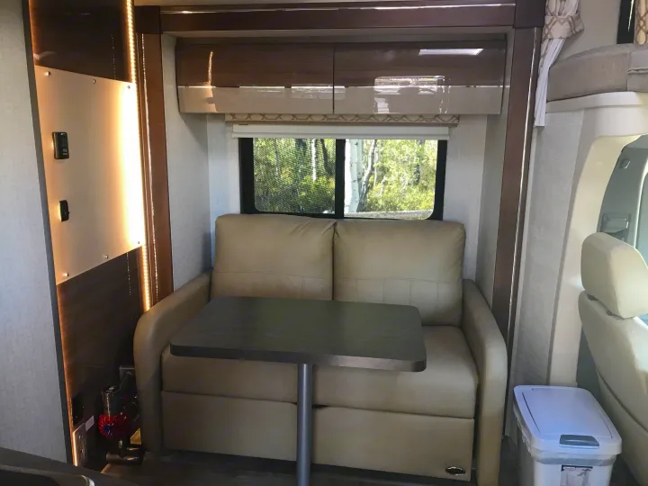 This rv rental's interior features a tan sofa with a dining table, ample lighting, and a window showcasing a natural view.. Itasca Navion 2017