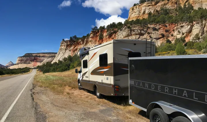 Scenic RV rental adventure: A Winnebago Navion and trailer parked roadside by towering, layered rock cliffs on a sunny day.. Itasca Navion 2017