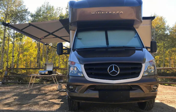 Winnebago rv rental, a Mercedes-Benz chassis, parked with awning extended. Grill ready amidst autumn trees for camping.. Itasca Navion 2017