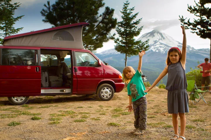 Family fun on an RV rental trip! Kids wave by their red camper van with a mountain view, perfect for outdoor adventures.. Volkswagen Eurovan weekender 2003