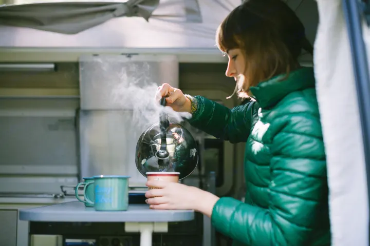 Woman in green jacket pouring steaming water from kettle into mug in an RV rental. Perfect for cozy camp mornings.. Volkswagen Eurovan camper 1999