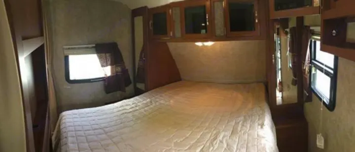 RV rental bedroom showing a bed with quilted comforter, overhead wood cabinets, and side windows.. Keystone Passport 2014