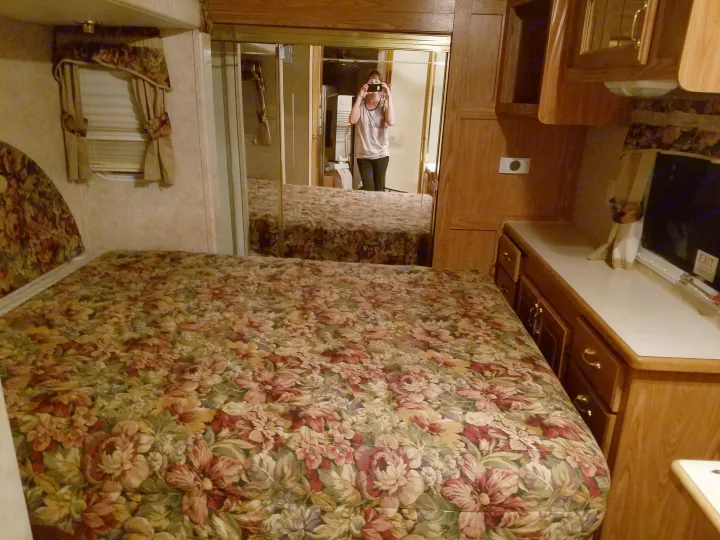RV rental bedroom featuring a floral bed, mirrored closet, window, and wood storage. Person reflected taking photo.. Forest River Cedar Creek Silverback Edition 2005