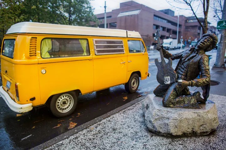 A bright yellow vintage RV rental is parked next to the iconic Jimi Hendrix statue on a wet urban street.. Volkswagen Westfalia 1977