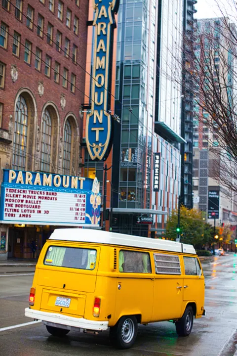 A yellow VW camper van, like an RV rental, drives past the iconic Paramount Theatre marquee in Seattle on a wet street.. Volkswagen Westfalia 1977