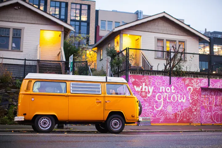 An orange vintage camper van, perfect for an RV rental, parked roadside. Behind it, glowing houses and a "why not grow?" mural.. Volkswagen Westfalia 1977