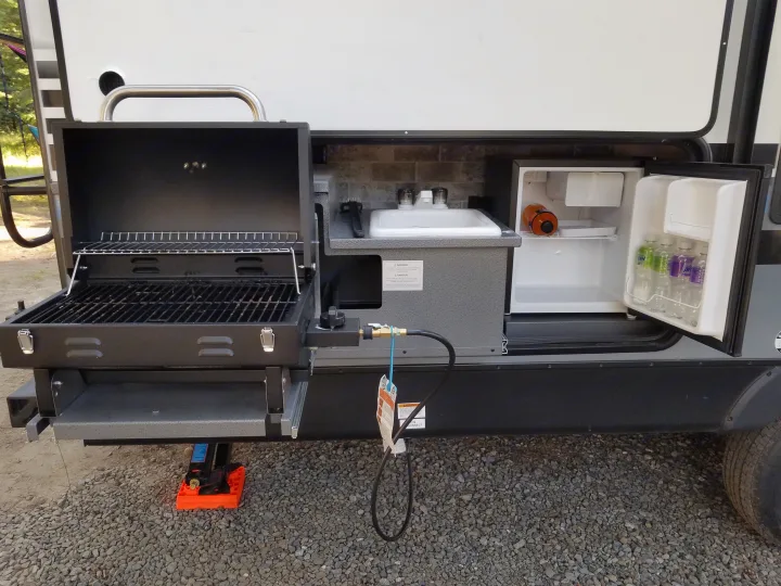 This RV rental's outdoor kitchen setup includes a BBQ grill, sink with faucet, and a mini-fridge stocked with drinks.. Forest River Surveyor 2018