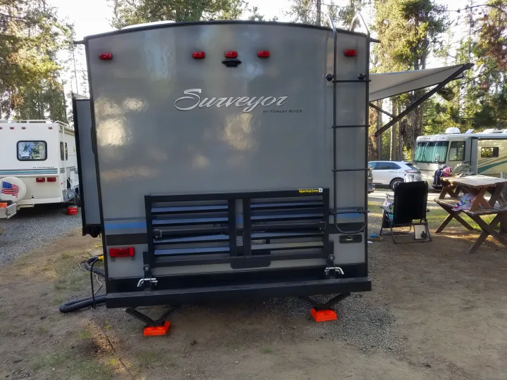Rear of a grey RV (Surveyor travel trailer) with storage rack, ladder, and orange leveling blocks, parked at an RV site for rv rental.. Forest River Surveyor 2018