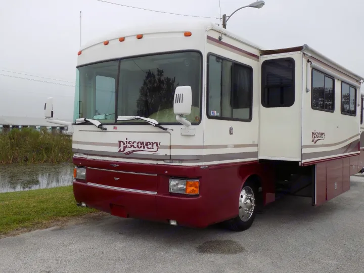 Front view of a white & maroon Discovery motorhome RV rental, parked with a slide-out extended near water.. Fleetwood Discovery 1998