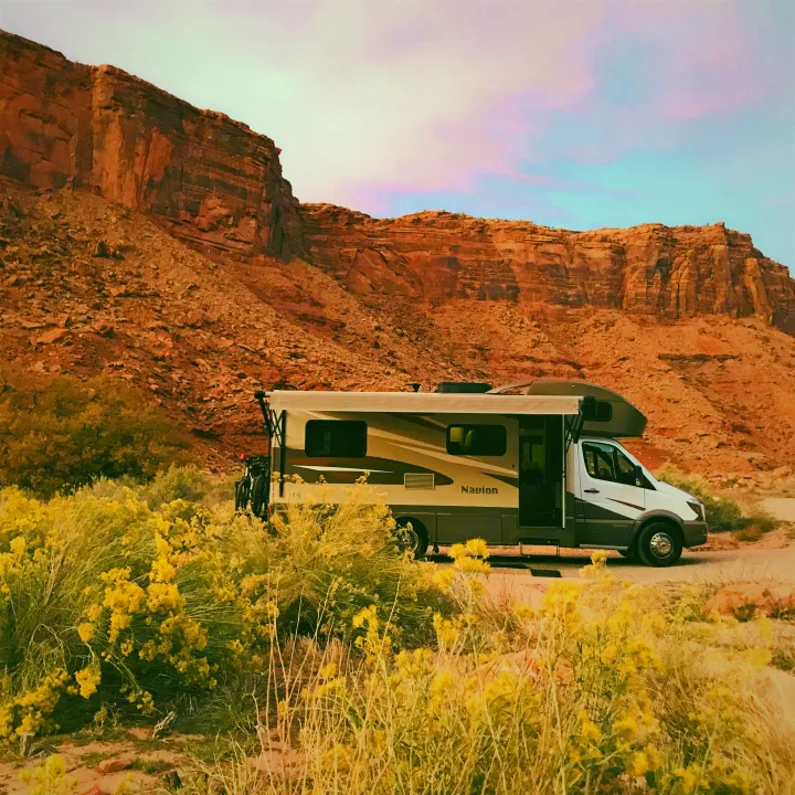 An RV rental is parked among yellow desert bushes, facing majestic red rock cliffs under a vibrant, pastel sky.. Itasca Navion 2017