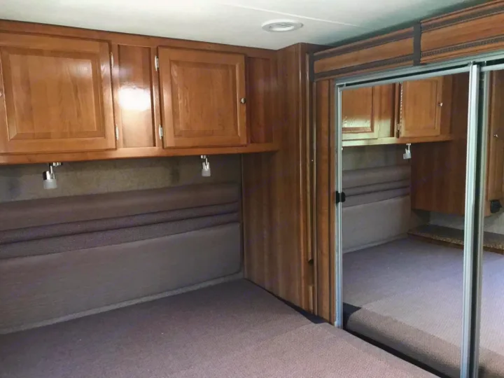 Cozy RV rental bedroom with wooden cabinets, reading lights, and a mirrored closet reflecting the compact sleeping area.. Coachmen Leprechaun 2018