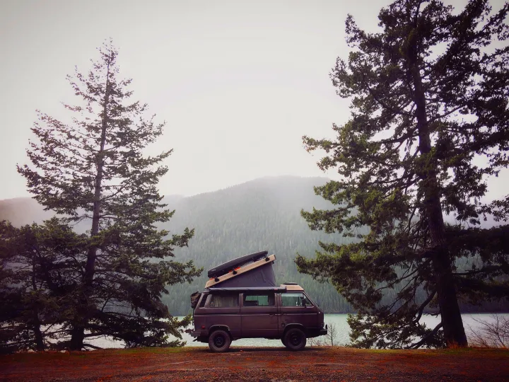 A dark camper van with pop-top, perfect for rv rental, parked on dirt by a misty lake with tall trees & forested mountains.. Volkswagen Vanagon 1985