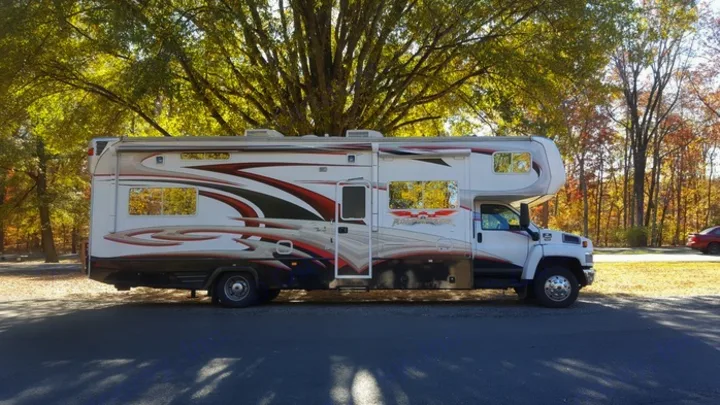 This striking RV rental motorhome, white with red graphics, is parked on asphalt amid bright autumn trees on a sunny day.. Weekend Warrior Weekend Warrior Motorhome 2009