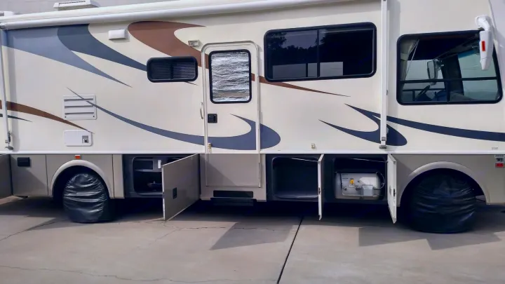 Cream RV rental with open storage bays and tire covers, parked on concrete. Ready for your next outdoor adventure.. National Surf Side 2006