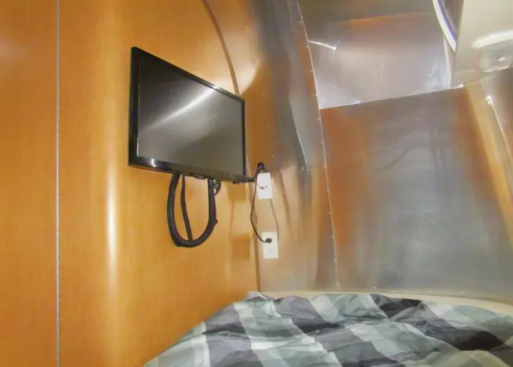 Inside an RV rental: a TV mounted above a bed with plaid covers, next to curved aluminum walls, ideal for travel.. Airstream Sport 2015