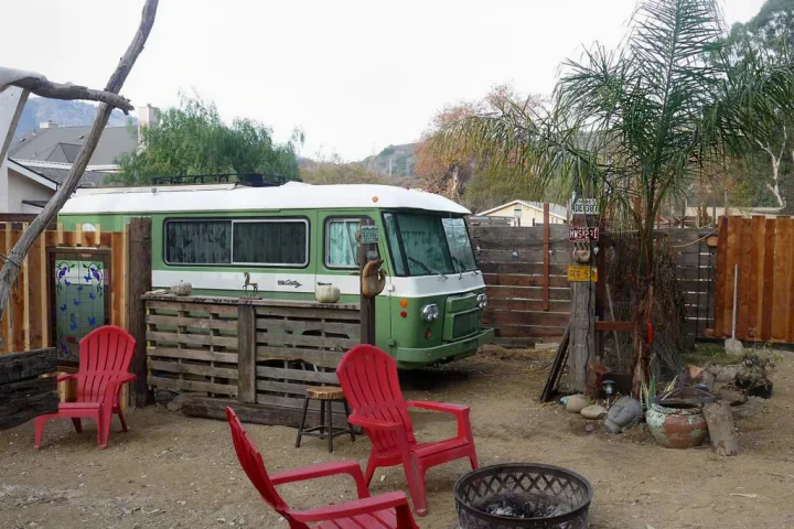 A unique green and white vintage bus RV rental setup with a pallet bar, red chairs, and a fire pit creates a rustic outdoor oasis.. Clark Cortez 1966