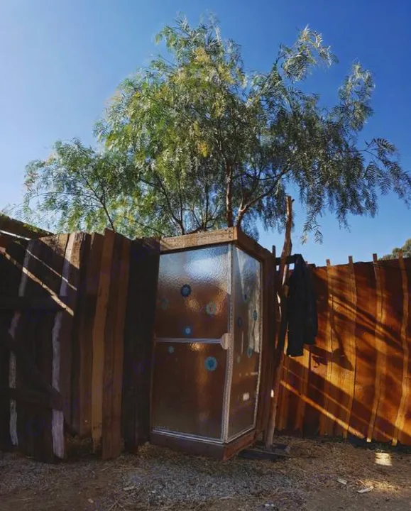 Rustic outdoor shower setup at an RV rental site. Wooden enclosure & clear panels under a tree, with a coat hanging nearby.. Clark Cortez 1966
