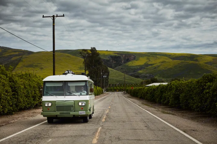 Green and white vintage RV rental on a scenic road with rolling yellow-green hills and orchards under cloudy skies.. Clark Cortez 1966