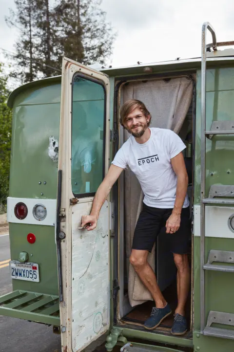 Smiling man standing in the doorway of a unique green vintage RV, ready for your next RV rental adventure.. Clark Cortez 1966