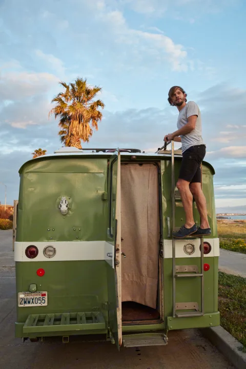 Exploring an RV rental: A man climbs a ladder on a vintage green camper, ready for adventure by the California coast.. Clark Cortez 1966