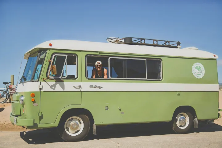 A man looks out from a vintage green and white RV rental, a Cortez camper van parked by sand on a sunny day.. Clark Cortez 1966