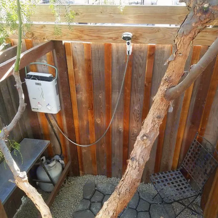 Outdoor shower setup for RV rental: portable water heater, shower head mounted on wooden fence, with propane tank. Gravel ground.. Clark Cortez 1966