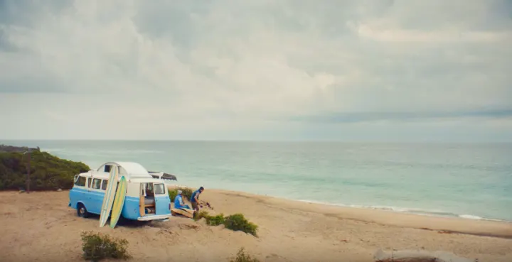 A light blue camper van, perfect for RV rental, on a sandy beach with surfboards. Two people relax by the ocean.. Ford Econoline 1966