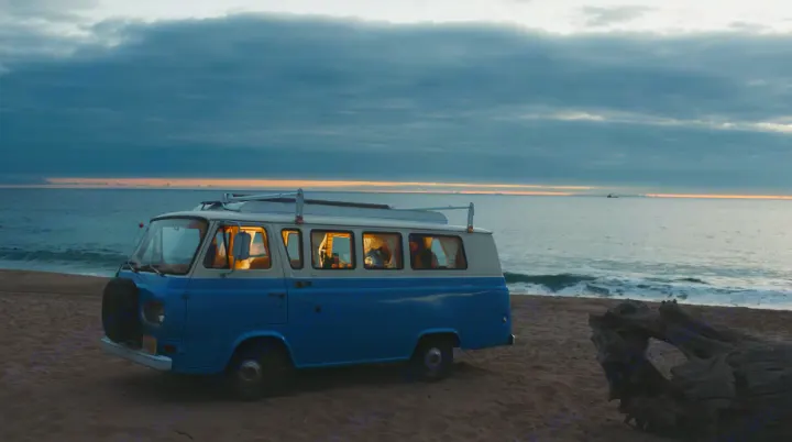 Vintage blue & white RV rental on a sandy beach at dusk, ocean in background. People inside the lit van.. Ford Econoline 1966