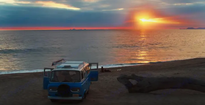 A classic RV rental sits on a sandy beach at sunset, facing the ocean. Golden light reflects on the water.. Ford Econoline 1966