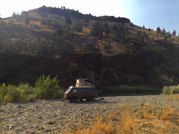 A brown camper van with pop-top tent parked on a rocky riverbank, mountains in background. Perfect for your RV rental adventure.. Volkswagen Vanagon 1985