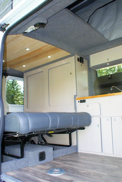 Inside this RV rental: modern camper van interior with a multi-purpose grey bench seat, wood ceiling, and compact kitchen.. Dodge Promaster 2017