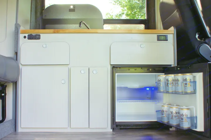 RV rental kitchen: light-toned cabinets, wood countertop with sink, outlets, and a fridge glowing blue, full of Modelo beer.. Dodge Promaster 2017
