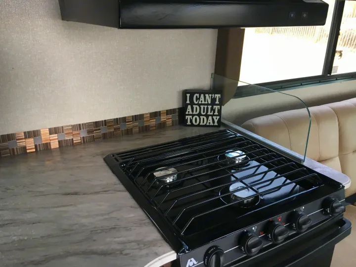 An RV rental kitchen, featuring a gas stovetop with a clear glass guard, tiled backsplash, and a playful "I CAN'T ADULT TODAY" sign.. Coachmen Freelander 2018