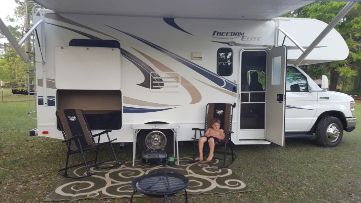 Family enjoying their RV rental: a Freedom Elite motorhome parked at a grassy campsite with a child, chairs, and grill.. Thor Motor Coach Freedom Elite 2015