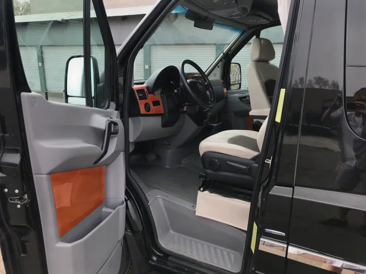 Inside a black RV rental van: Driver's seat with cream upholstery, wood-look dashboard, and open door.. Winnebago Era 2014