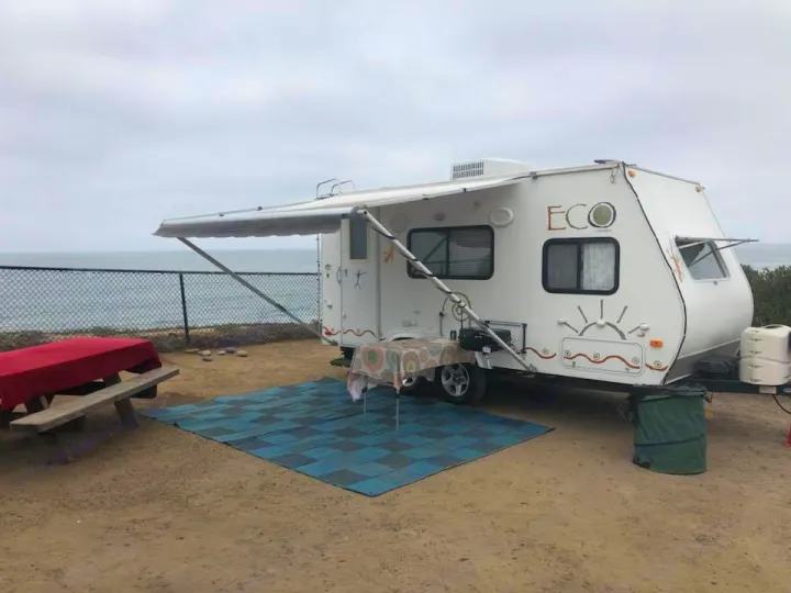 White ECO rv rental travel trailer parked oceanfront. Awning, table, blue mat on dirt campsite with a sea view.. Dutchman Eco 2010