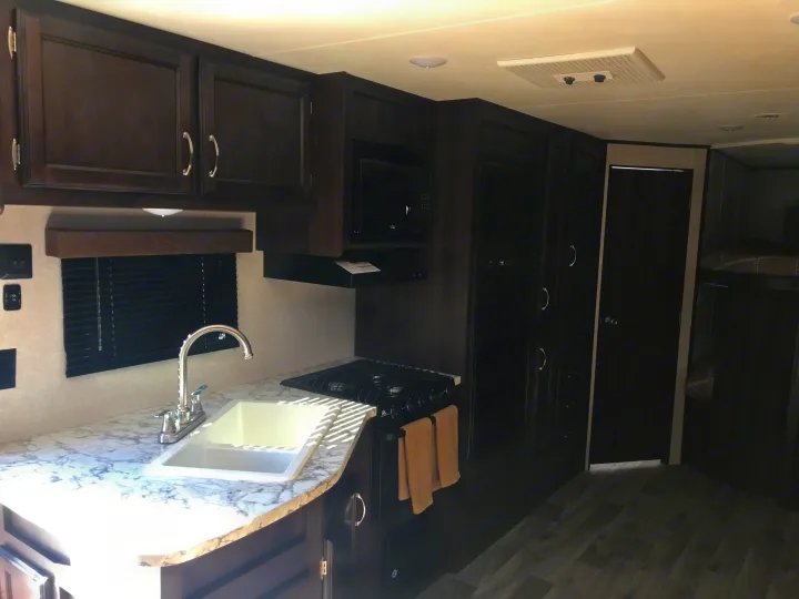 Modern rv rental kitchen showcasing dark cabinetry, white marble countertops, sink, stove, microwave, and a glimpse of bunk beds.. Jayco JAY FLIGHT SLX 287 BHS 2018