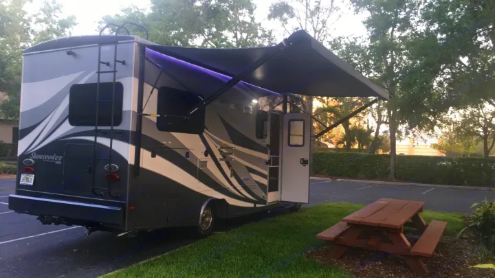 Modern rv rental with an extended awning and purple LED lights, parked at a lush campsite. Ideal for your next road trip adventure!. ForestRiver Sunseeker 2018