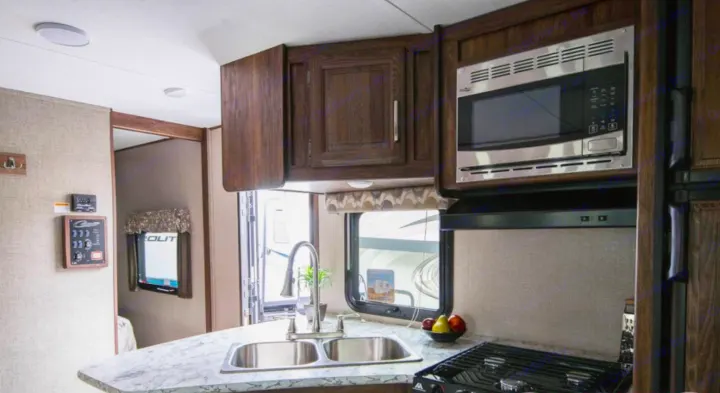 Modern RV rental kitchen featuring a stainless steel microwave, double sink, and gas stovetop amidst wood cabinetry.. Keystone Cougar Half-Ton 2018