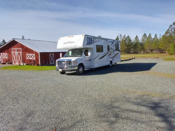 White Sunseeker RV rental parked on a gravel lot beside a red barn and tall trees, ready for your next adventure.. Forest River Sunseeker 2013