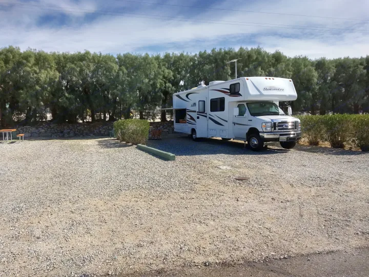 White Sunseeker rv rental on gravel lot. Dog, picnic table, and green trees under a partly cloudy sky.. Forest River Sunseeker 2013