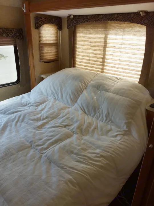 Inside an RV rental: a bright bedroom with a fluffy white comforter, sunlit windows with blinds, and wooden accents.. Forest River Sunseeker 2013