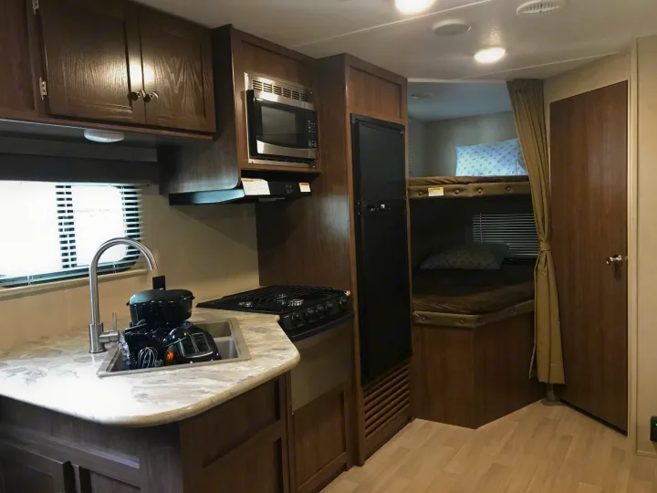 Inside an rv rental, showing a compact kitchen area with sink, stove, microwave, and fridge, next to bunk beds.. Coleman Lantern 2018