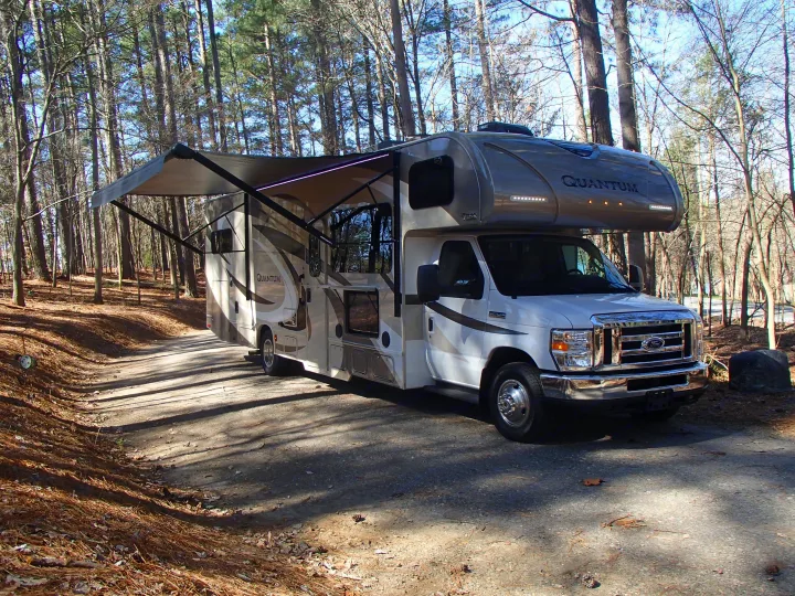 White Thor Quantum RV rental with an extended awning parked in a sunny, tree-filled campsite, ideal for outdoor adventures.. Thor Quantum 2017