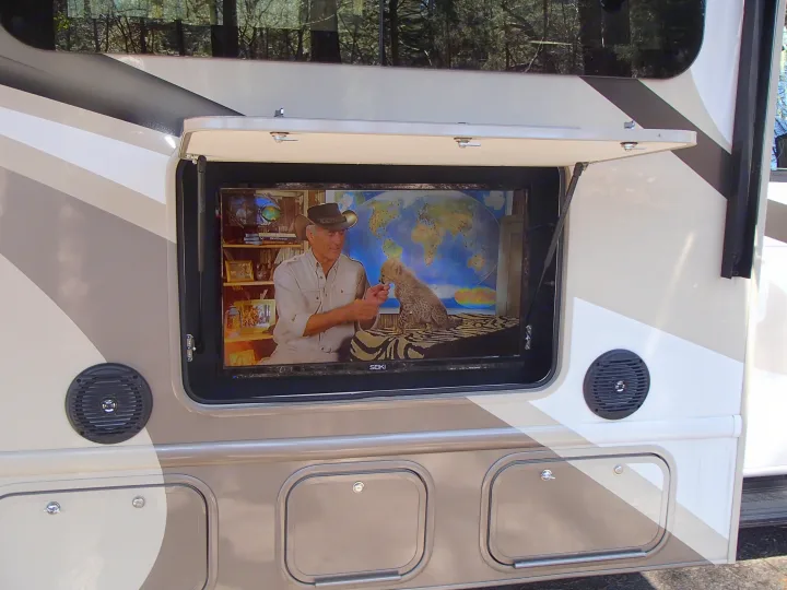 An RV rental's outdoor TV is open, displaying a man with a baby cheetah and a world map. Speakers are below it.. Thor Quantum 2017