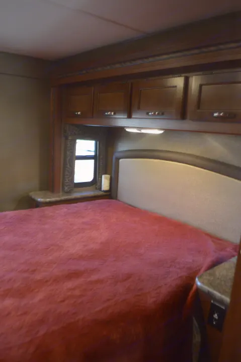Cozy RV rental bedroom features a red-blanketed bed, dark wood cabinets, and a side window.. Thor Motor Coach Chateau 29G 2016
